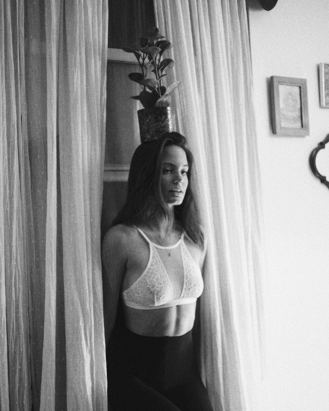 Mujer con camiseta de encaje y pantalones oscuros, de pie junto a cortinas en una habitación. victoria secret, boudoir editorial 