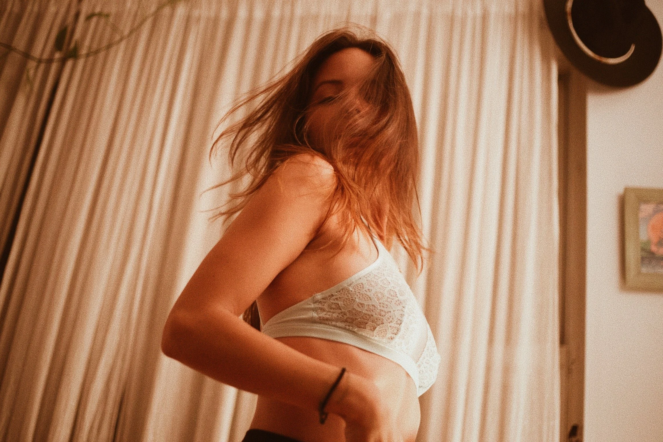 Mujer con cabello largo y castaño claro en un cuarto, con cortinas beige de fondo, usando ropa interior blanca de encaje