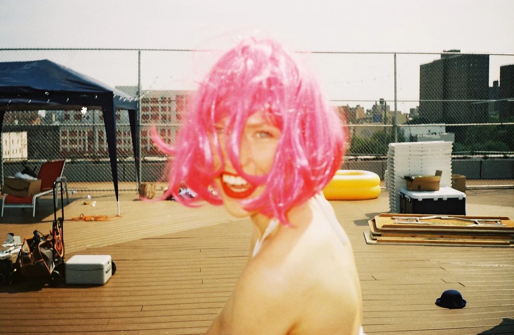 Persona con cabello rosa, sonriente y usando labial rojo en una terraza al aire libre con objetos y estructuras en el fondo.
