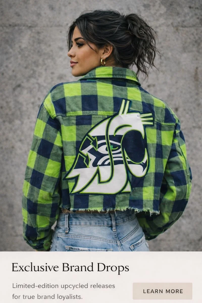 A woman with dark hair in a messy bun, wearing a Custom green and navy plaid oversized jacket with a large embroidered logo on the back, and light-wash jeans, standing against a gray concrete wall.