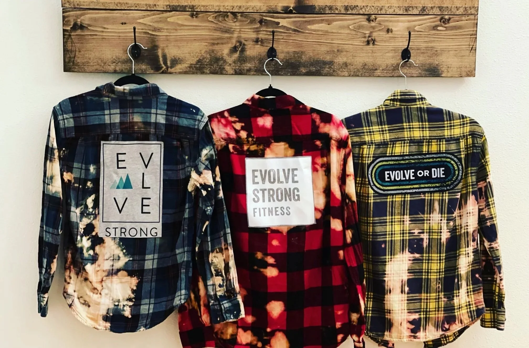 Three plaid shirts hanging on black hangers against a wooden wall. The left shirt is blue plaid with a label that reads 'Live Strong.' The middle shirt is red plaid with a label that says 'Evolve Strong Fitness.' The right shirt is yellow plaid with a label that reads 'Evolve or Die.'