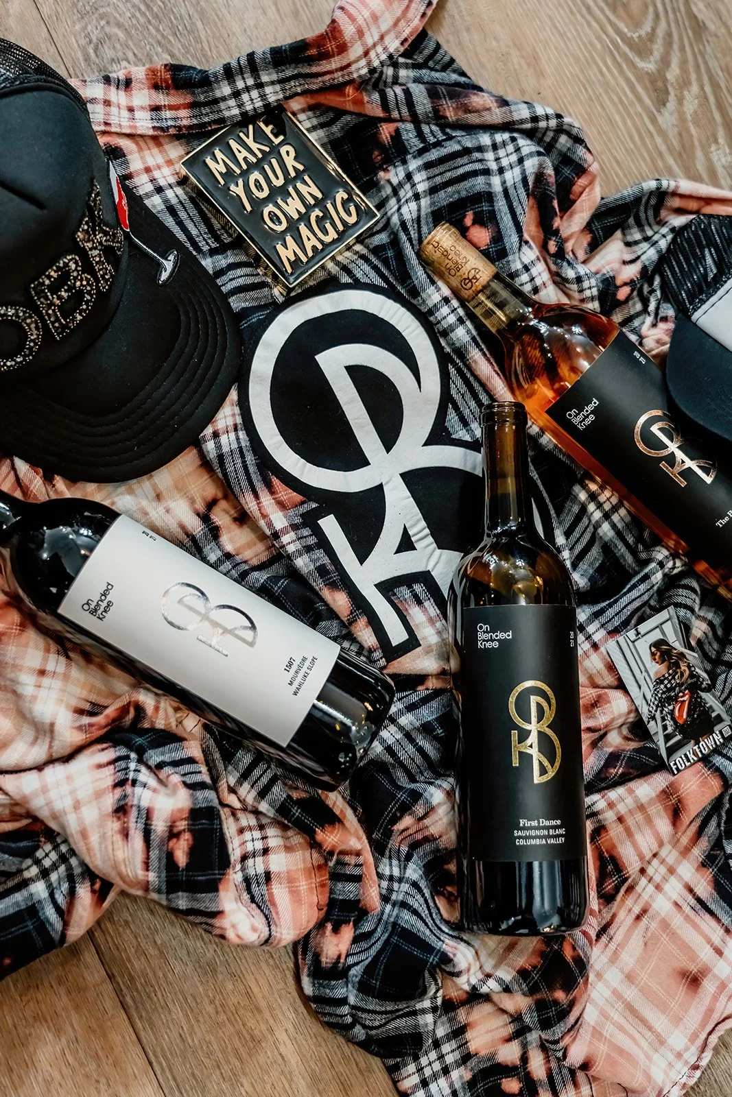 A collection of alcohol bottles, a black cap, a plaid shirt with a 'Make Your Own Magic' pin, a photo, and a large logo on fabric, all arranged on a wooden surface.