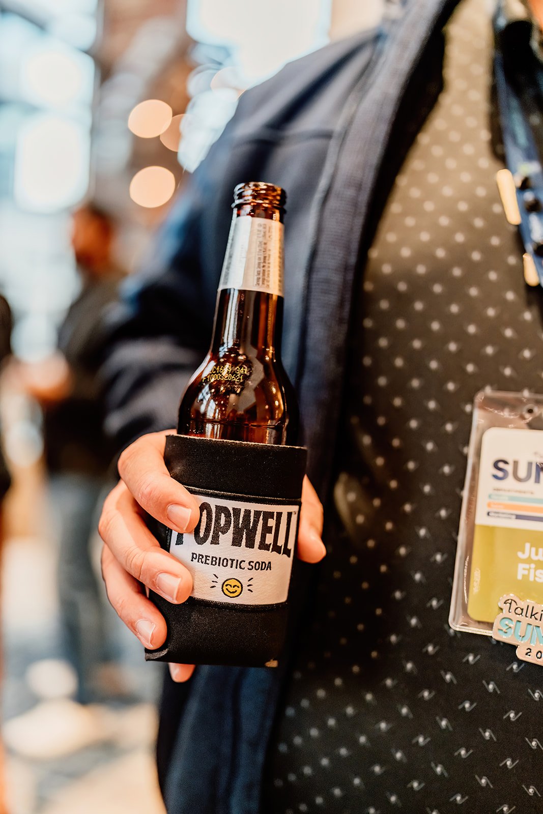 Person holding a bottled beverage in a black neoprene koozie labeled "Topwell Prebiotic Soda" with a smiling emoji, wearing a suit jacket with a name badge on a conference or event.