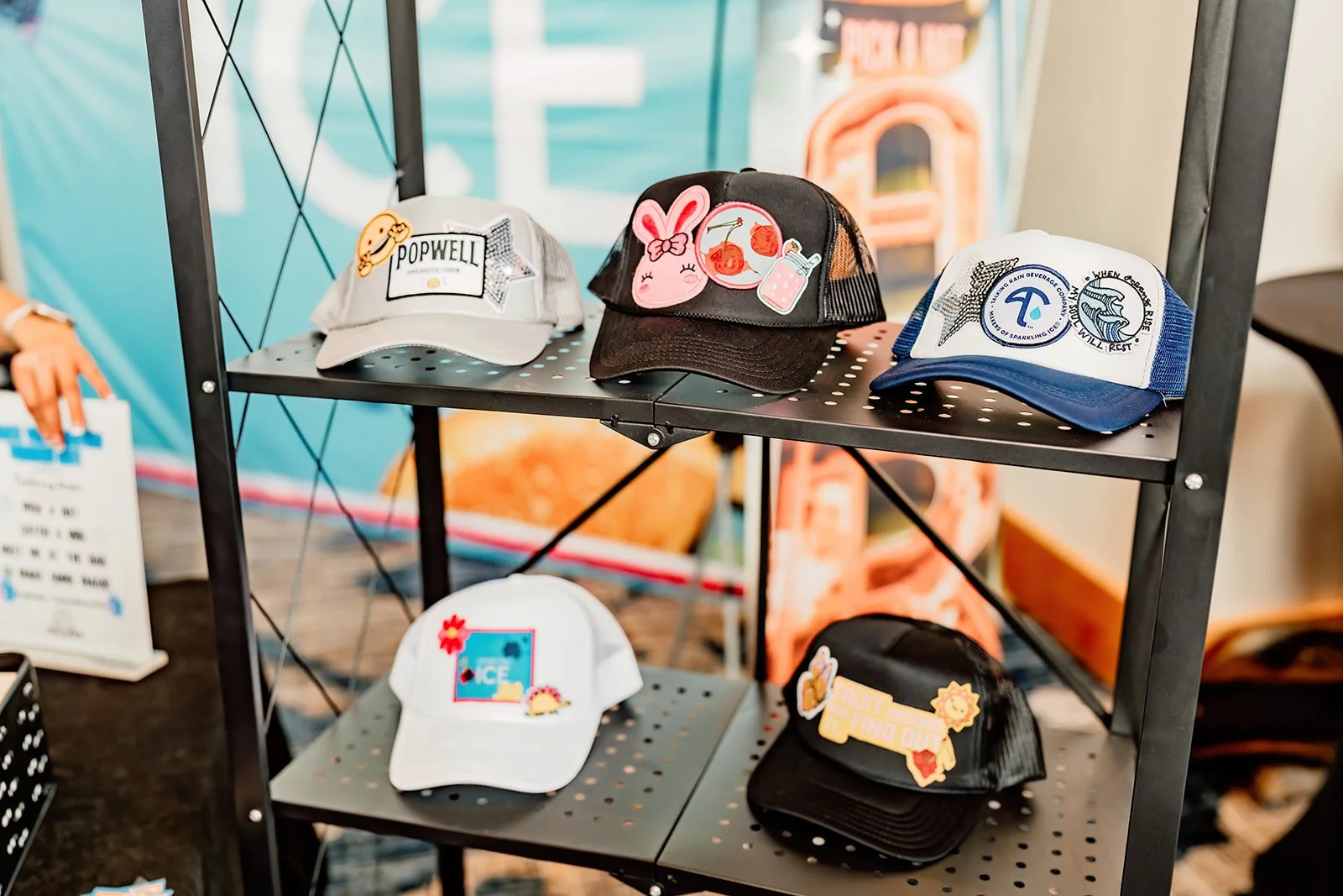 Display of six caps with various designs and patches on a black metal shelf at an indoor event.