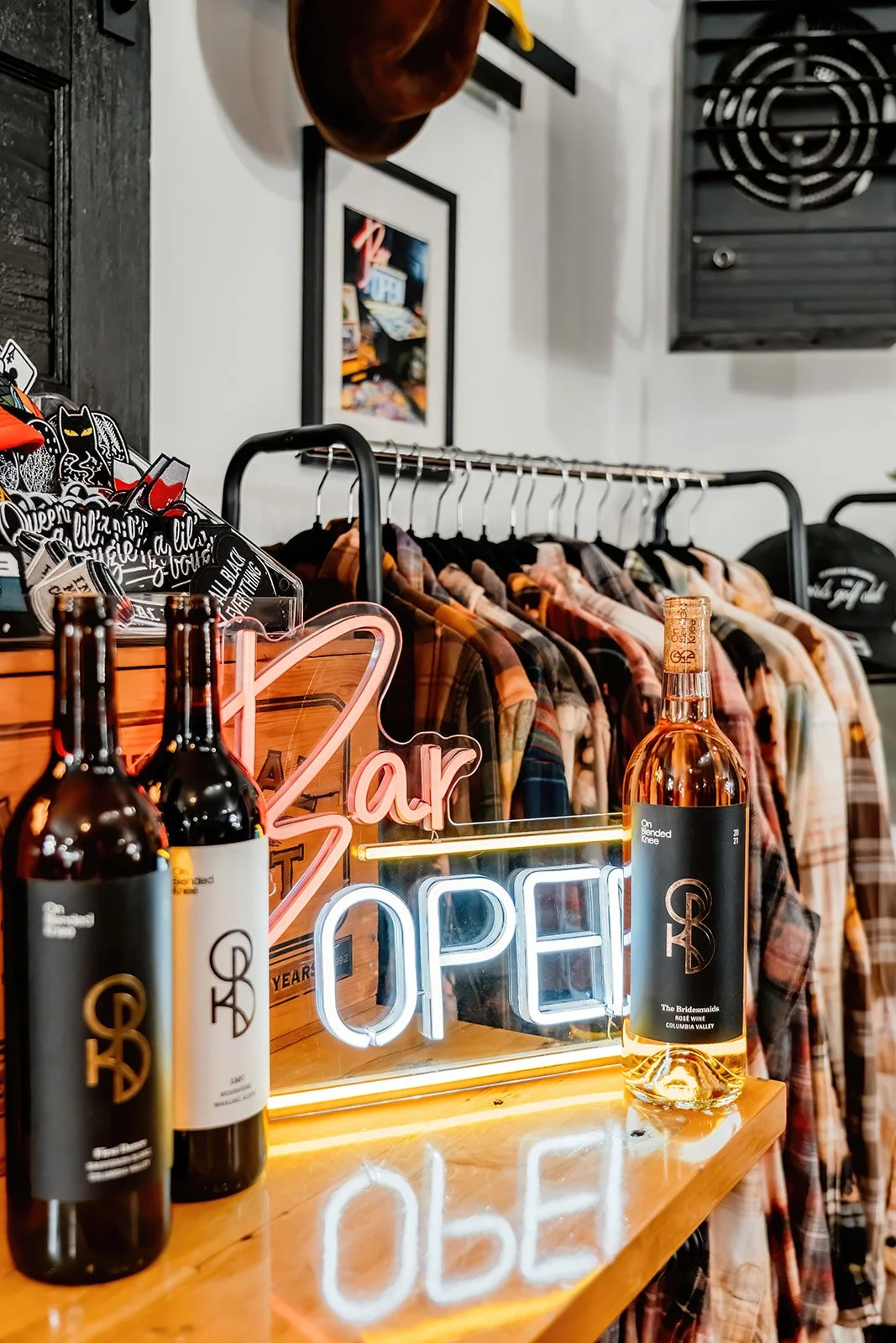 Neon sign reading 'Open' with bottles of rosé wine on a wooden table in front of a clothing rack with flannel shirts.