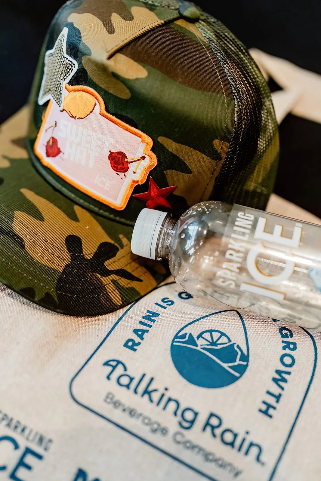 Camouflage baseball cap with patches, a clear water bottle labeled 'ICE,' and a white fabric bag with a blue logo that reads 'Walking Rain Beaverdge Conservancy'.
