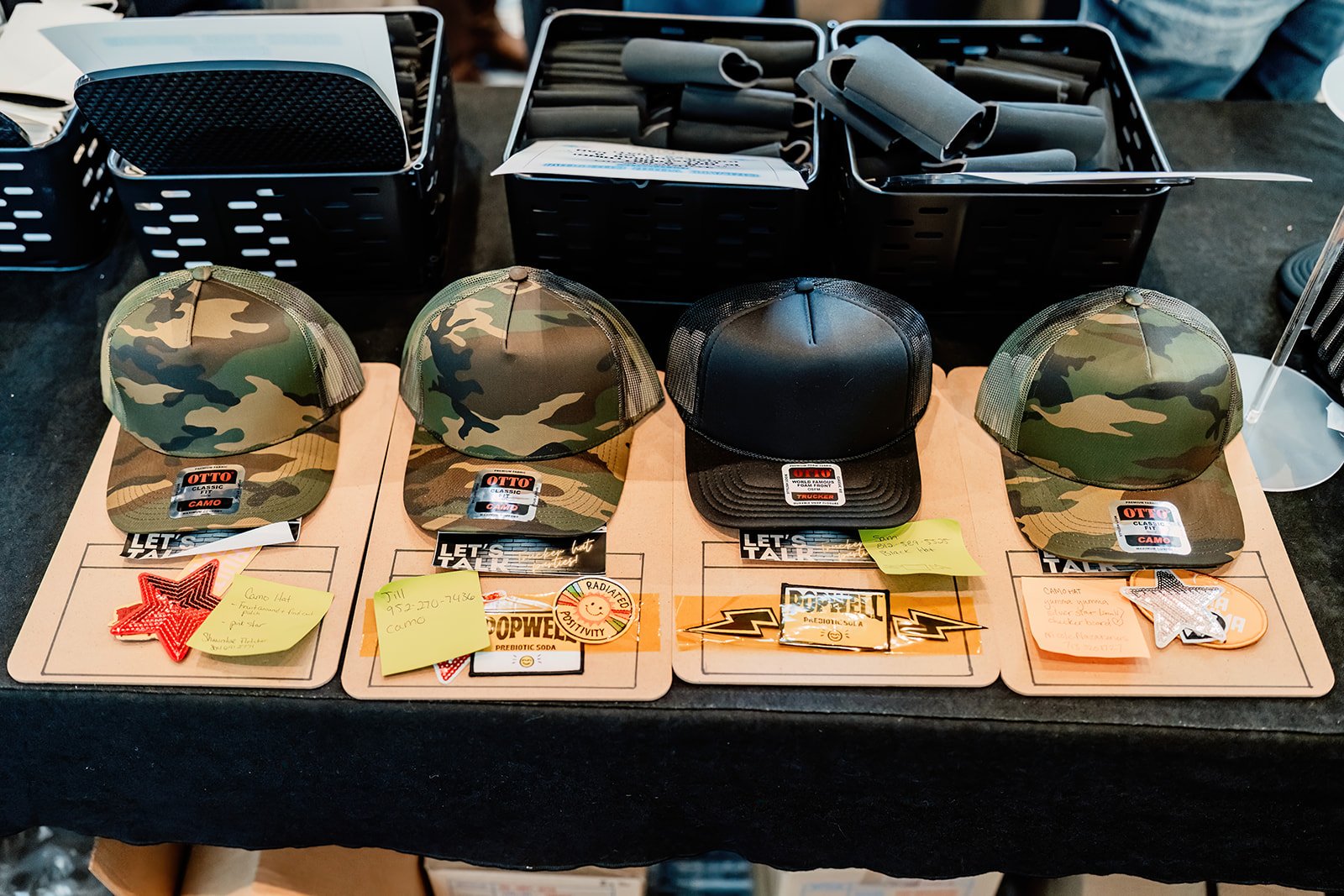 Four camouflage and one black baseball hats displayed on a table, each with stickers and notes underneath, with baskets of black tubes in the background.