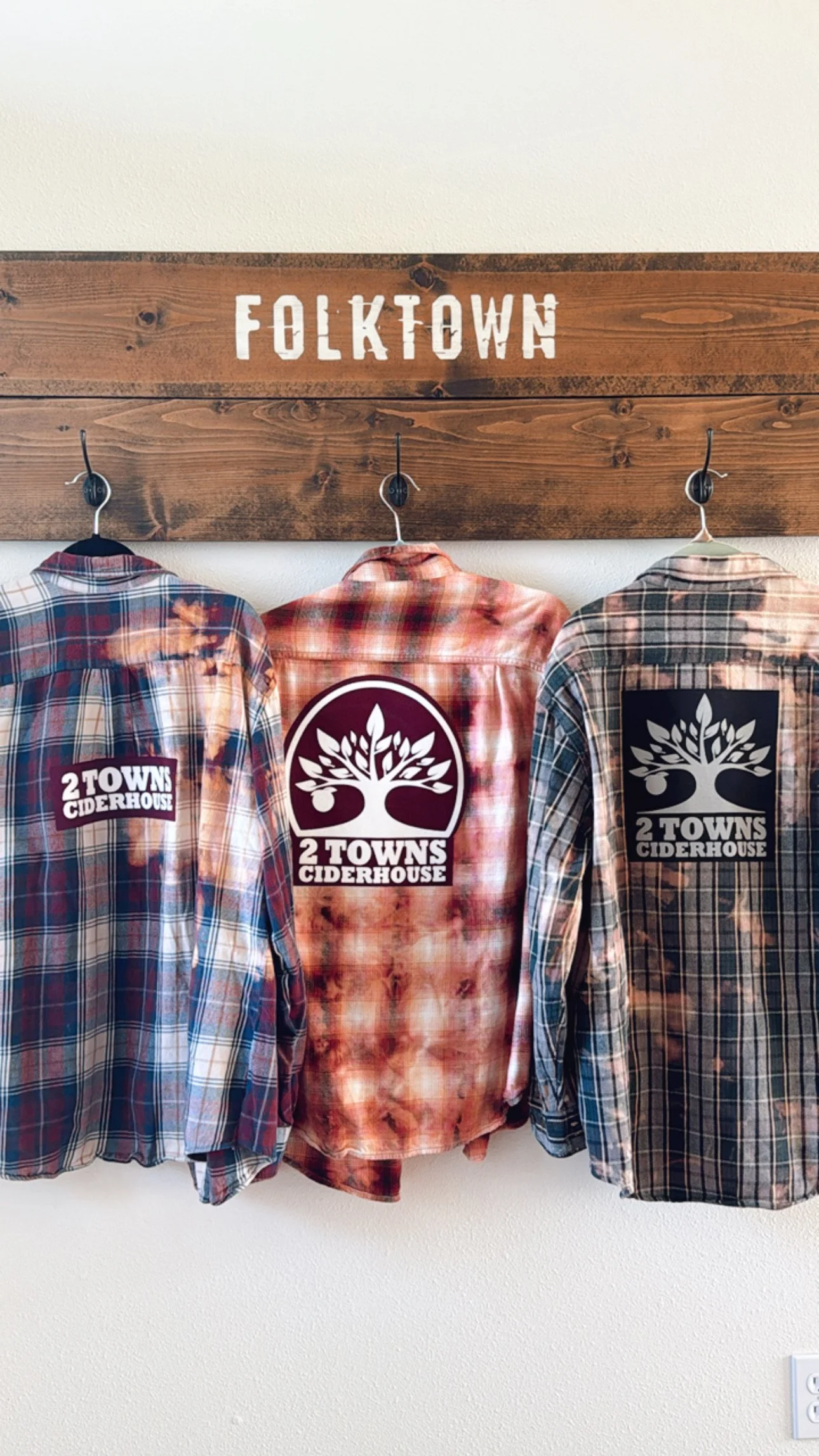 Three plaid shirts hanging on hooks under a wooden sign that reads 'FOLKTOWN,' with logos for 2 TOWNS CIDERHOUSE on the back of the shirts.