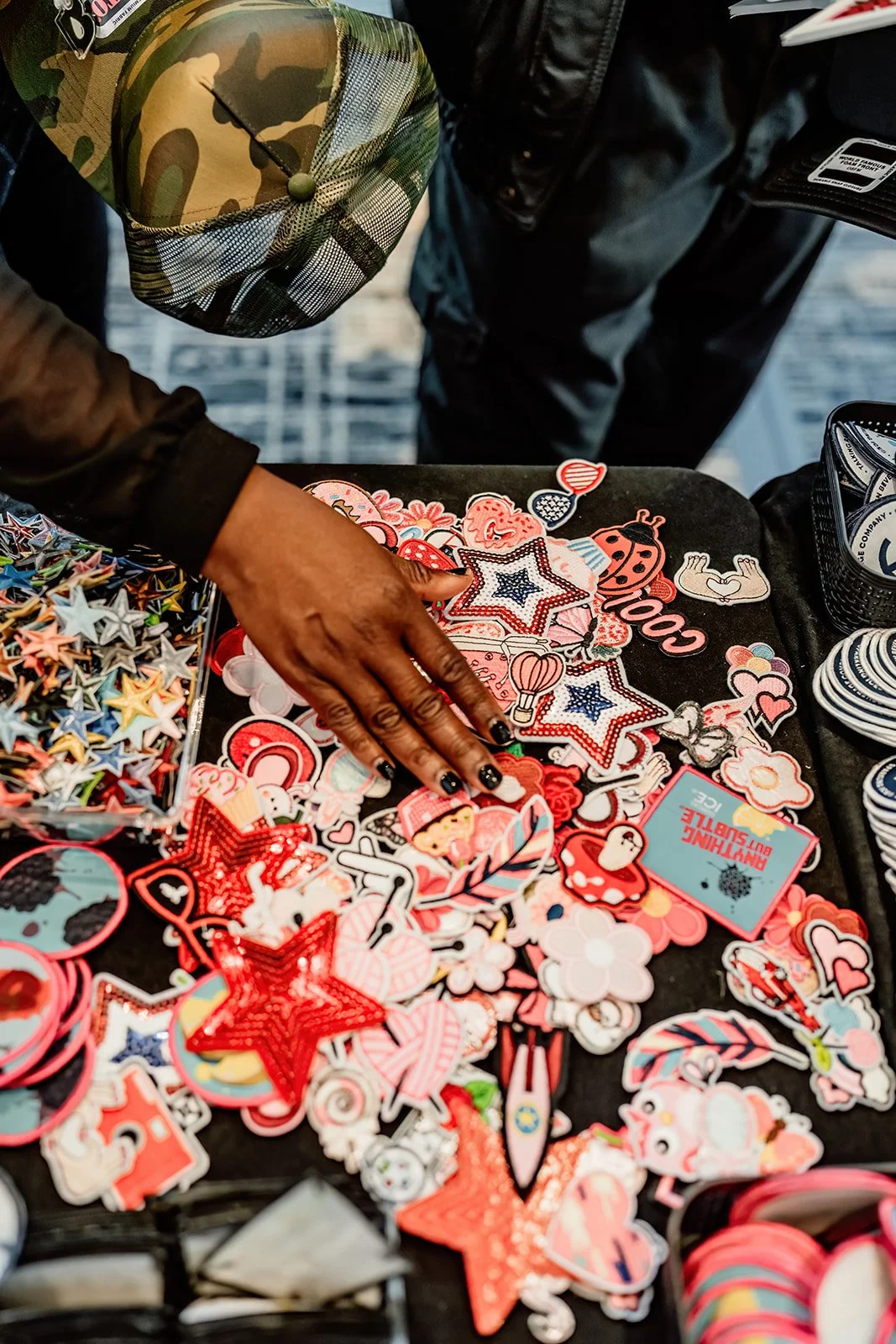 A person shopping for colorful patches and stickers at a street market stall. The patches include stars, hearts, flowers, and other playful designs, mostly in red, pink, and white. The person is wearing dark nail polish and a camouflage cap.