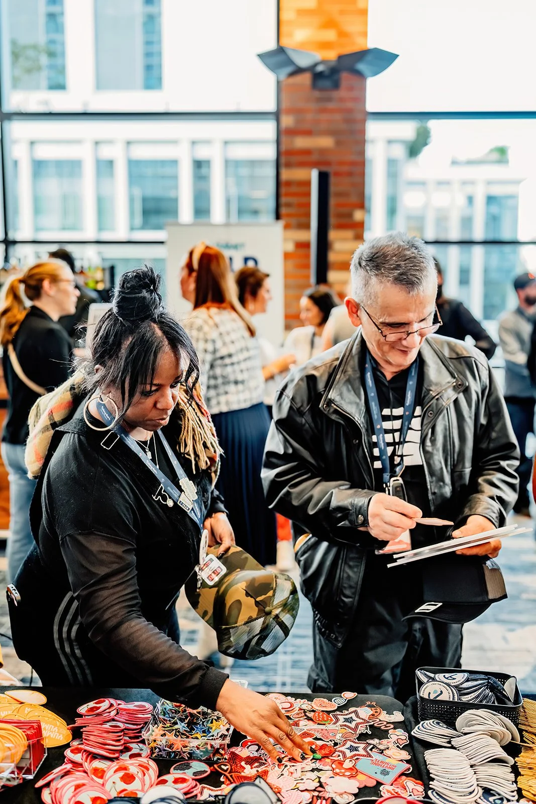 People browsing a table of red, white, and multicolored buttons and pins at an indoor event with large windows and a brick pillar.