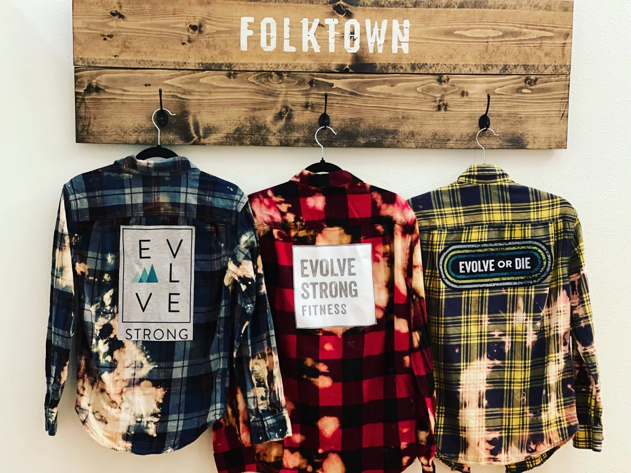Three plaid shirts hanging on hooks on a wooden sign that says 'FOLKTOWN' with phrases on the back of each shirt: left shirt says 'EVOLVE STRONG', middle shirt says 'EVOLVE STRONG FITNESS', right shirt says 'EVOLVE OR DIE'.