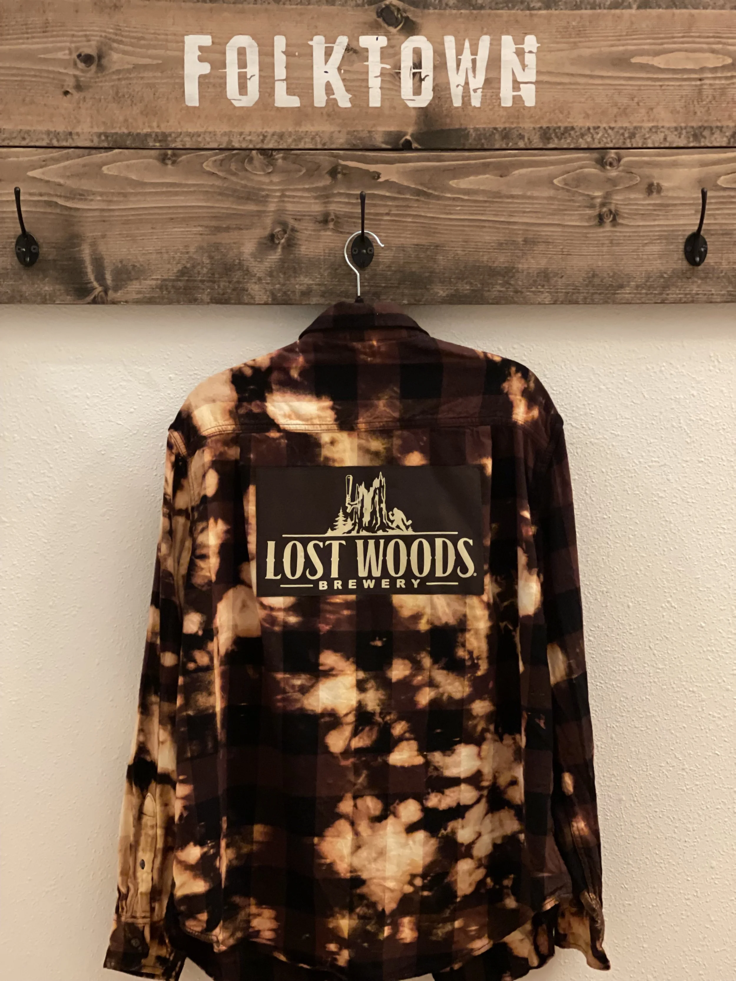 A plaid shirt with a 'Lost Woods Brewery' patch hanging on a wooden hook rack with 'FOLKTOWN' painted on top.