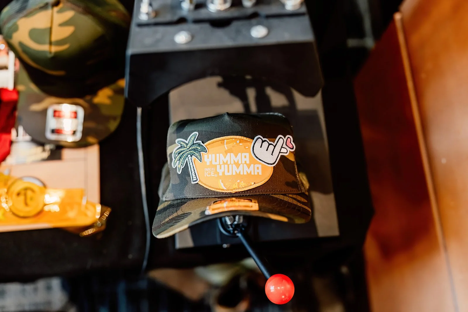 Camouflage baseball cap with patches that say 'Yumma Yumma', a palm tree, and a hand making a rock and roll sign with a heart, on a black surface.