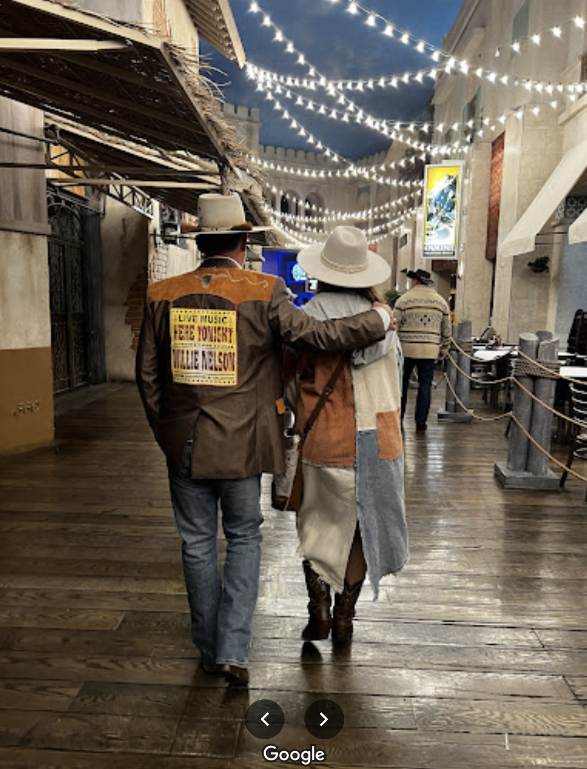 Two people walking arm-in-arm in an indoor shopping or entertainment area decorated with string lights hanging from the ceiling. One is wearing a cowboy hat, a patchwork jacket, and jeans, and the other is wearing a wide-brimmed hat, a jacket, and bo