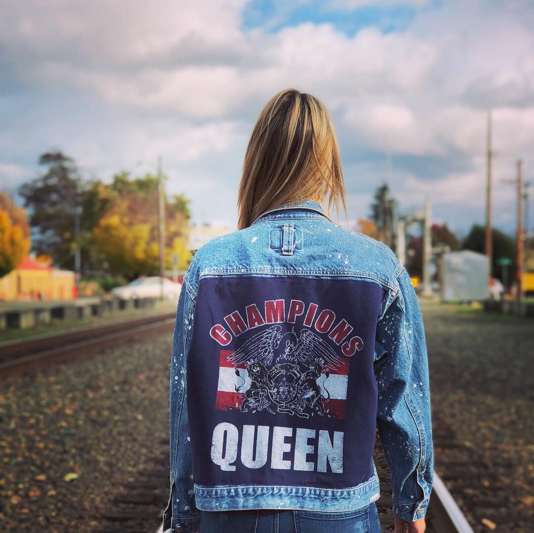 Woman wearing upcycled denim jacket with a patch that says 'CHAMPIONS QUEEN' and a graphic of a lion and eagle, standing near train tracks outdoors.