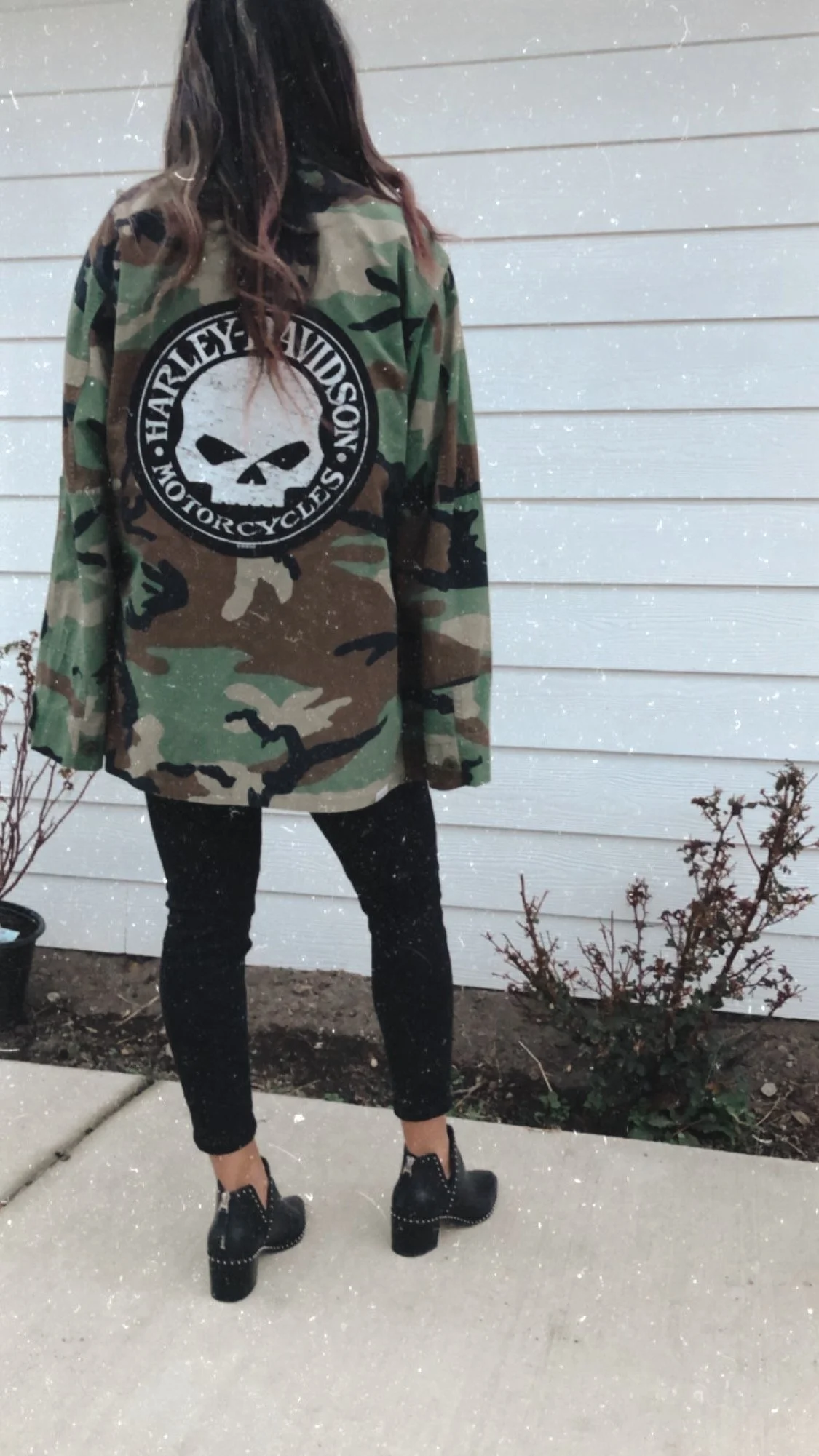 Person wearing a camouflage Harley-Davidson jacket, black pants, and black heeled boots, standing outdoors on concrete sidewalk in front of a white siding wall with small potted plants nearby.