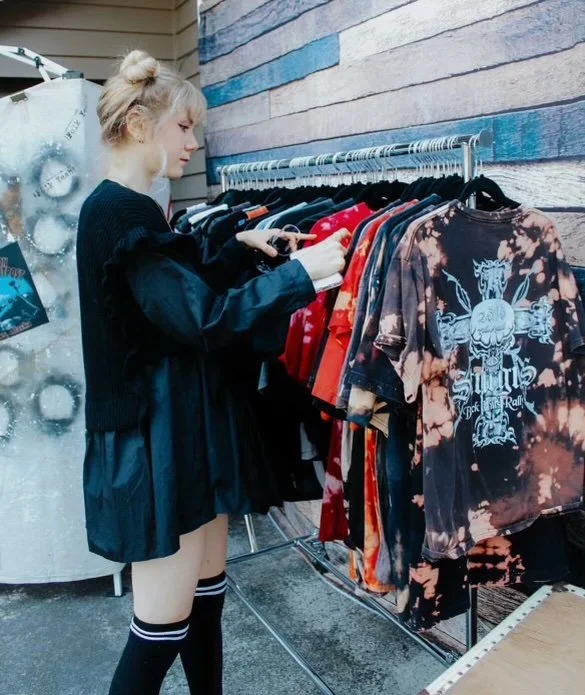 A young woman with blonde hair in a bun shopping for clothes on a metal rack outside. She is wearing a black jacket, shorts, and knee-high socks, and is browsing t-shirts, including a tie-dye design.