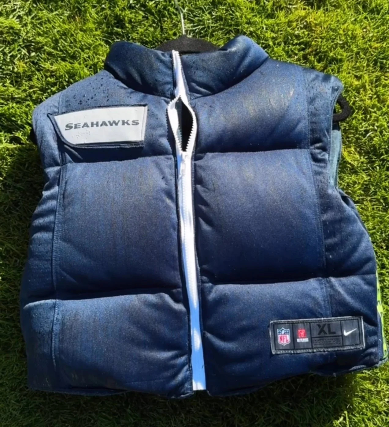 Blue sleeveless Custom Upcycled Seahawks jacket with NFL and Nike logos, and a patch indicating size XL, lying on grass.