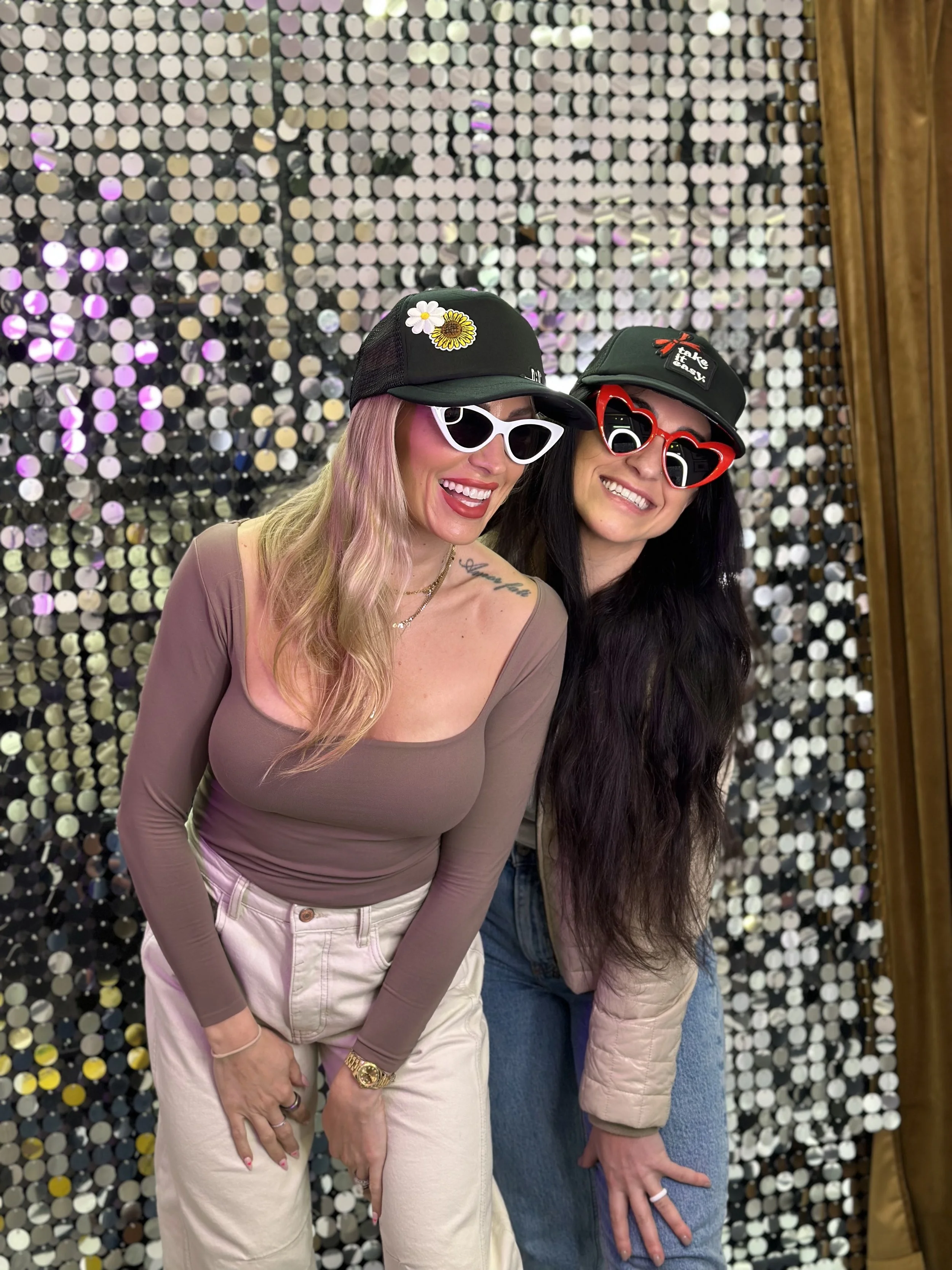 Two women smiling and posing for a photo in front of a shimmering sequin backdrop, wearing hats and colorful sunglasses.