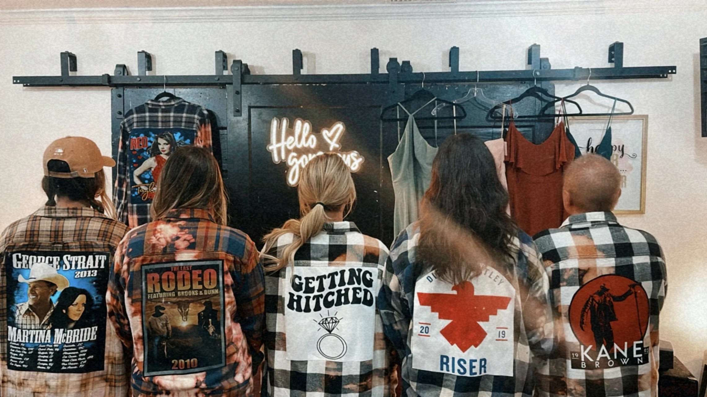 Five people standing with their backs facing camera, looking at a display of clothing and a neon sign that reads 'Hello Gorgeous' on a black wall.