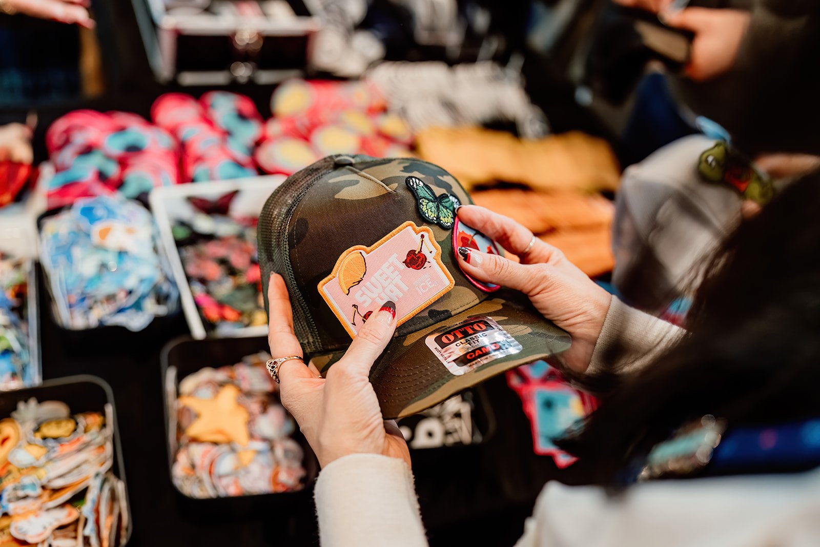 Person shopping at a stall with various colorful patches and accessories, holding a camouflage cap with patches and a butterfly pin.