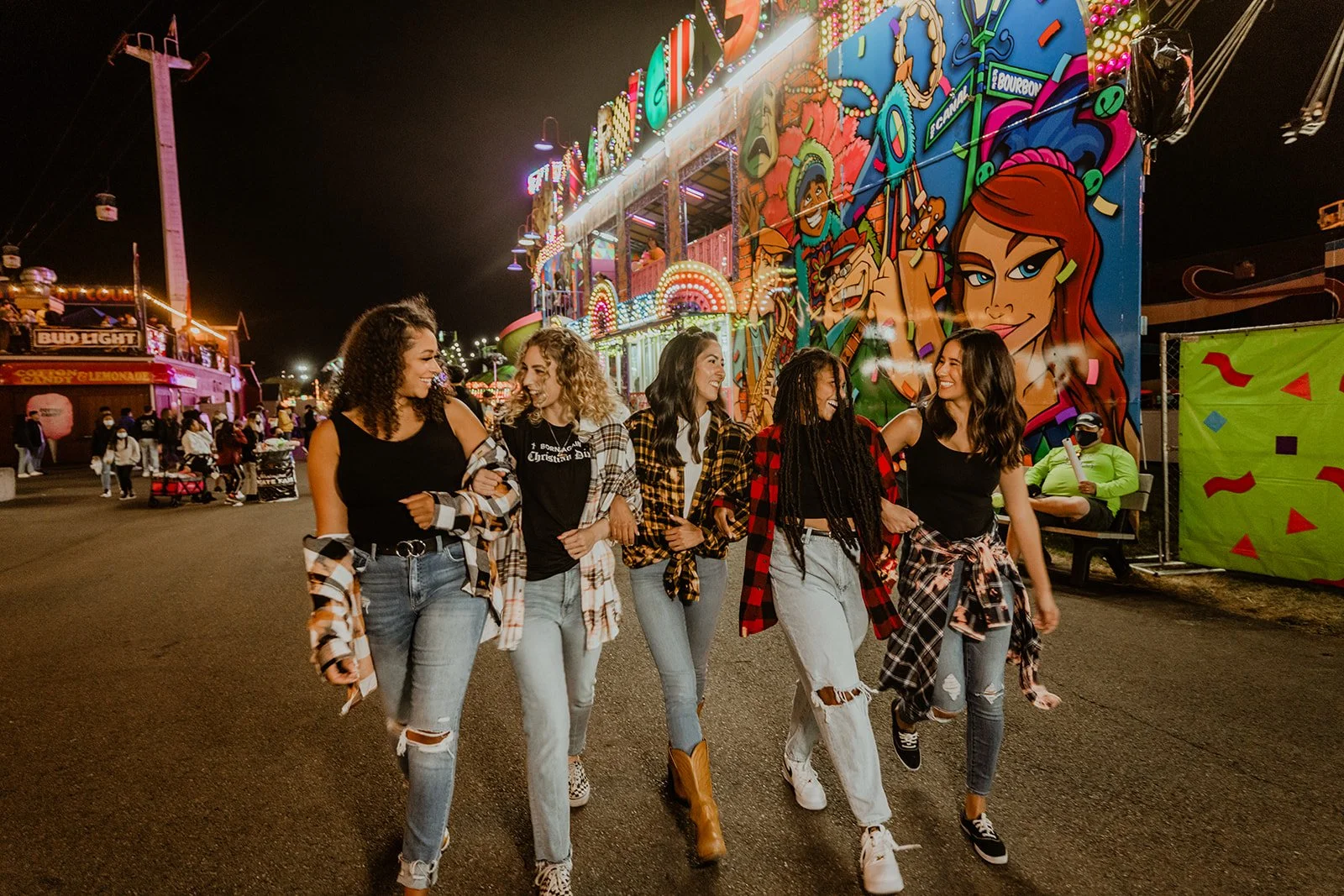 Group of six young women walking together at a carnival at night, with colorful lights and bright graffiti-style wall art in the background.