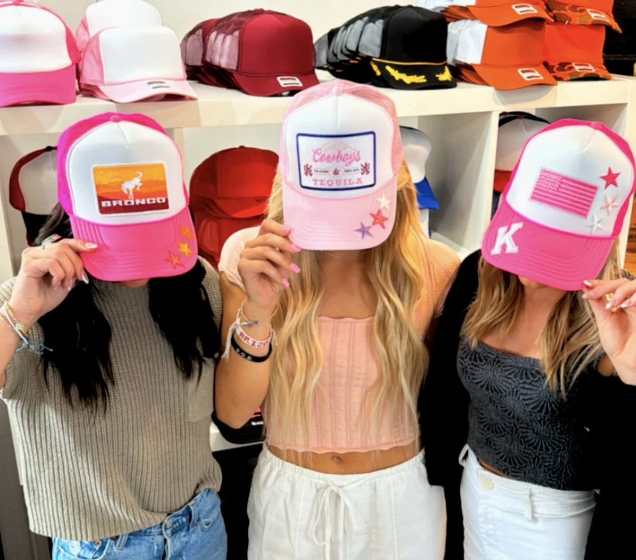 Three women in a store wearing baseball caps with custom patches, hiding their faces. The caps are pink, white, and pink with different designs and patches. Behind them are shelves with more caps in various colors.