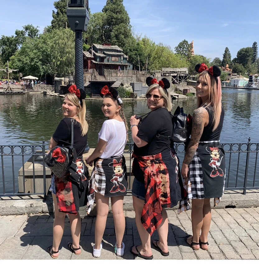 Four women are standing side by side by a water body, each wearing Disney-themed Minnie Mouse ears and matching skirts with Minnie Mouse designs. They are smiling and posing for the photo, with a scenic background featuring trees and a rustic buildin