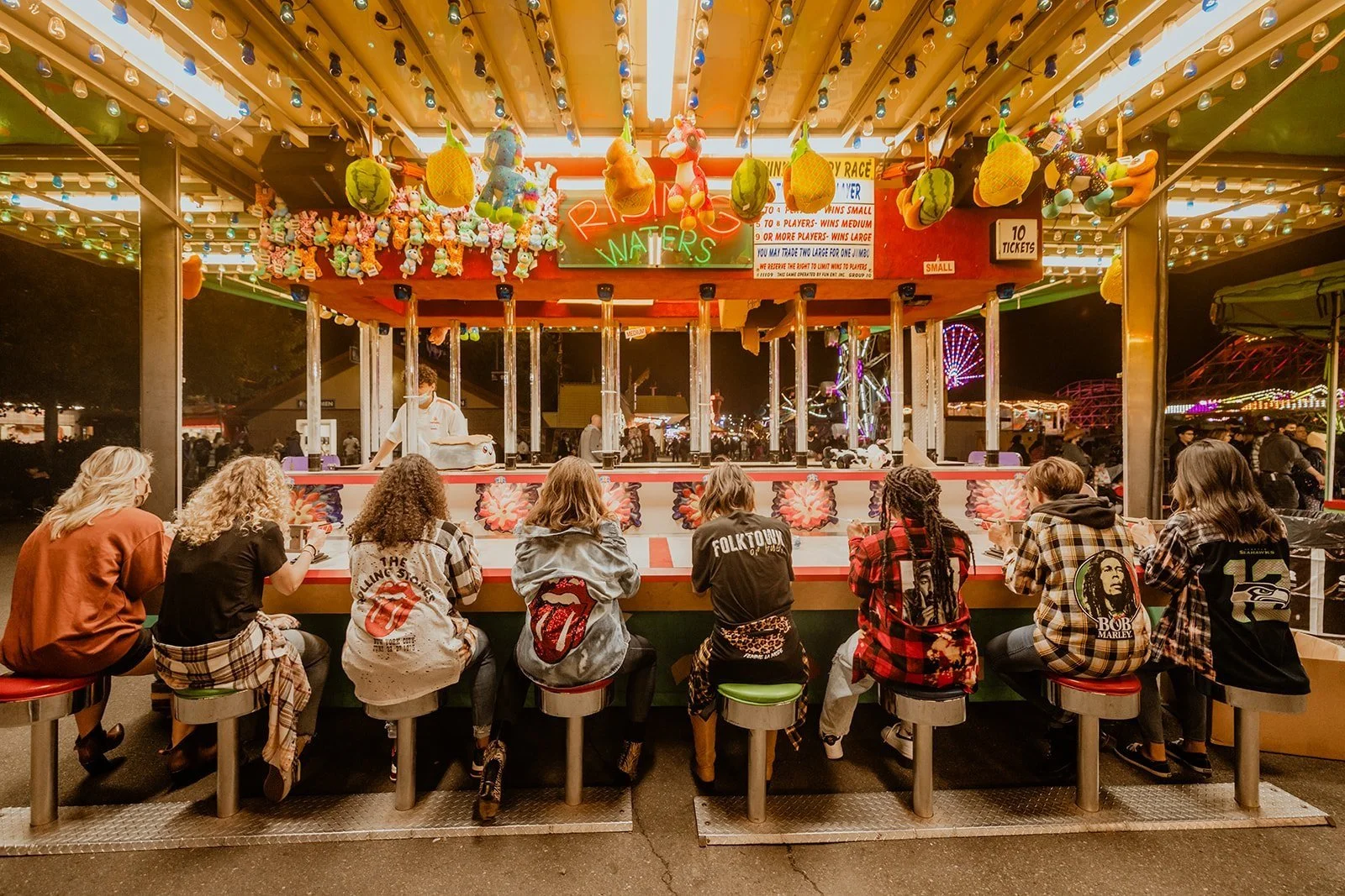A group of people sitting on stools at a carnival game booth, with plush toys hanging overhead and a brightly lit Ferris wheel in the background at night.