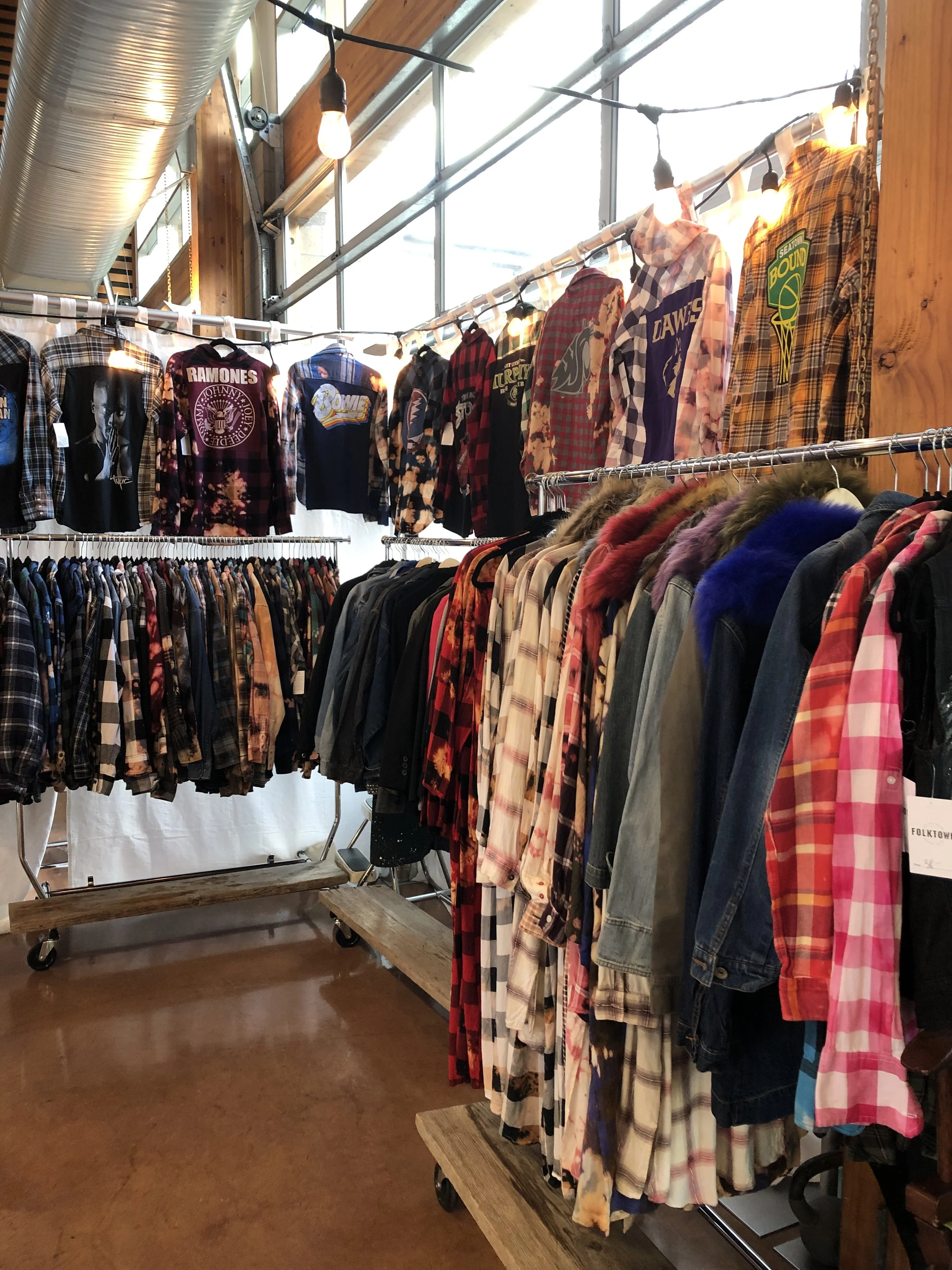 A clothing store display with racks of flannel shirts and jackets, and some shirts hanging on a higher rack, featuring various patterns and colors, illuminated by warm ceiling lights.