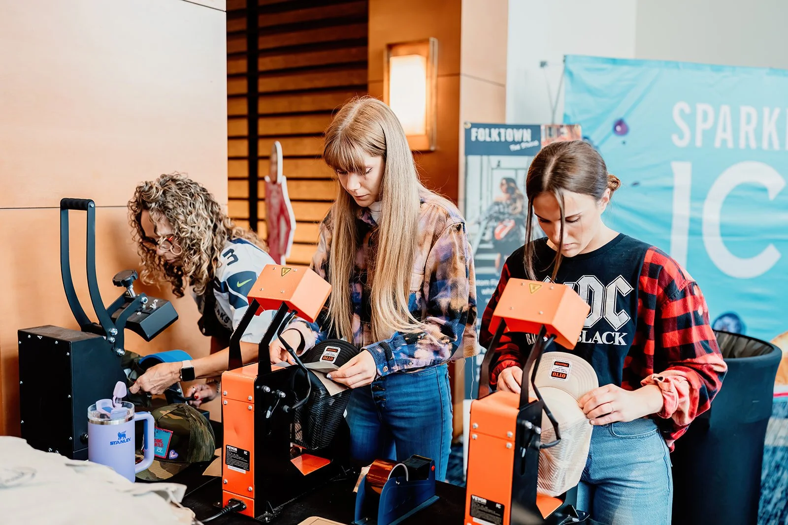 Three women at a booth with orange heat press machines, preparing to stencil or customize fabric items, in a setting with a wooden wall, a poster, and a large blue banner in the background.