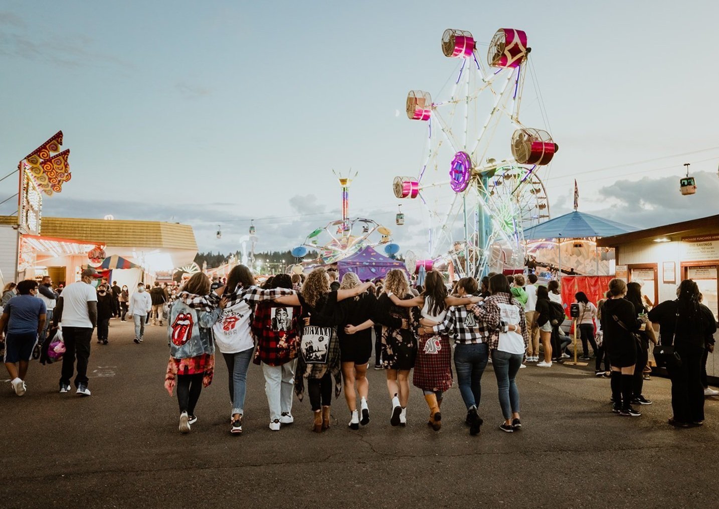Group of young women holding hands and walking together at a carnival, with amusement rides like a Ferris wheel and other attractions in the background, during evening time.