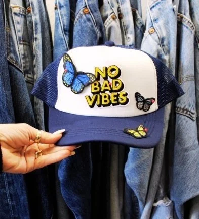Cap with a butterfly patch and the phrase 'No Bad Vibes' in yellow, held by a person against a denim jacket background.