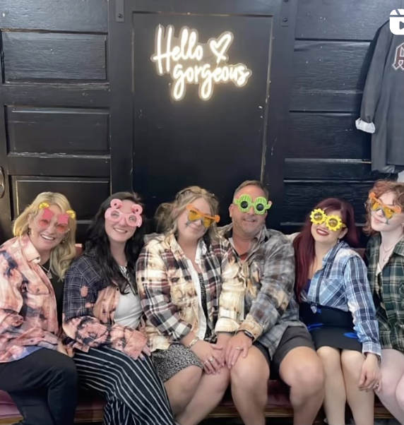 Group of six people sitting together, wearing colorful glasses, in front of a sign that says "Hello Gorgeous" illuminated in neon.