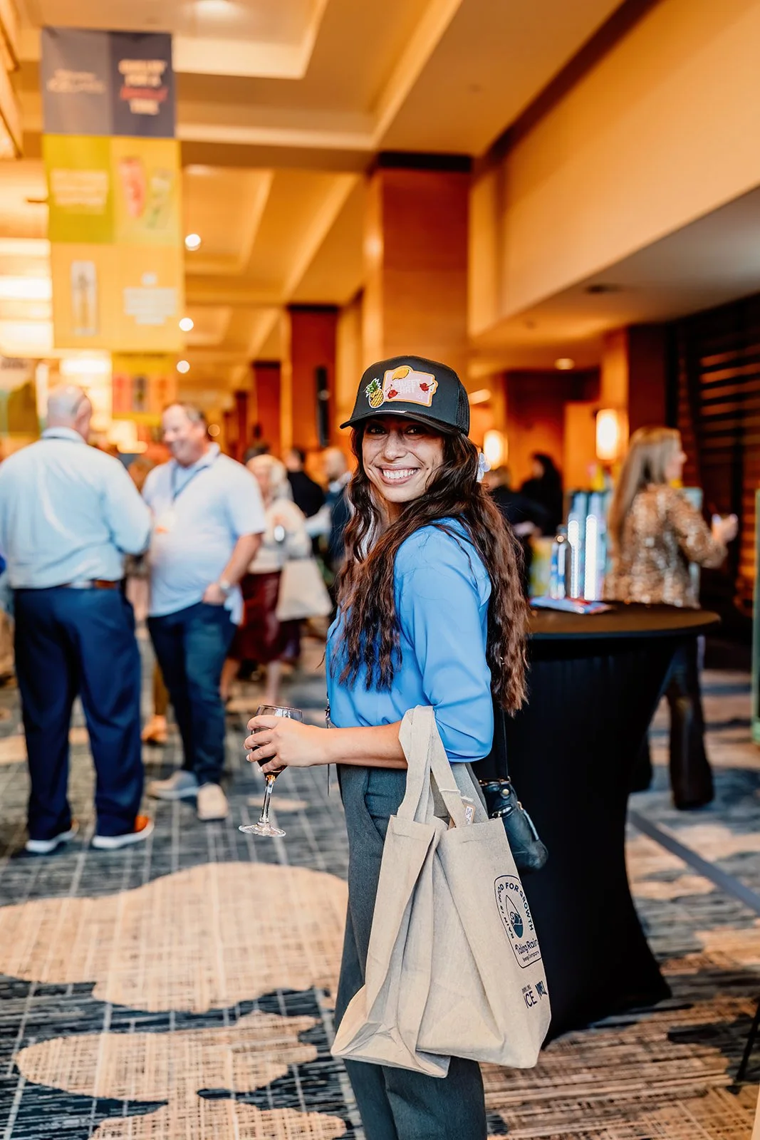 A woman with long wavy hair smiling and holding a wine glass, wearing a black cap with embroidered patches, a blue shirt, and gray pants, at a busy indoor event with people in the background.