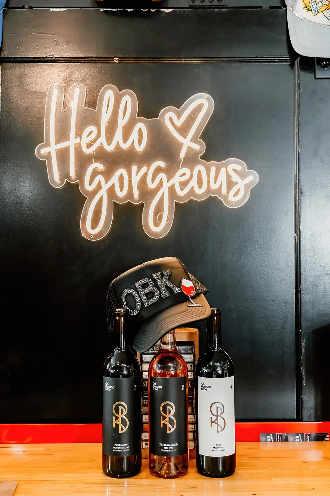 Decorative signs on a black wall with bottled wines and a baseball cap on a wooden surface. The signs say 'Hello gorgeous' with a heart.