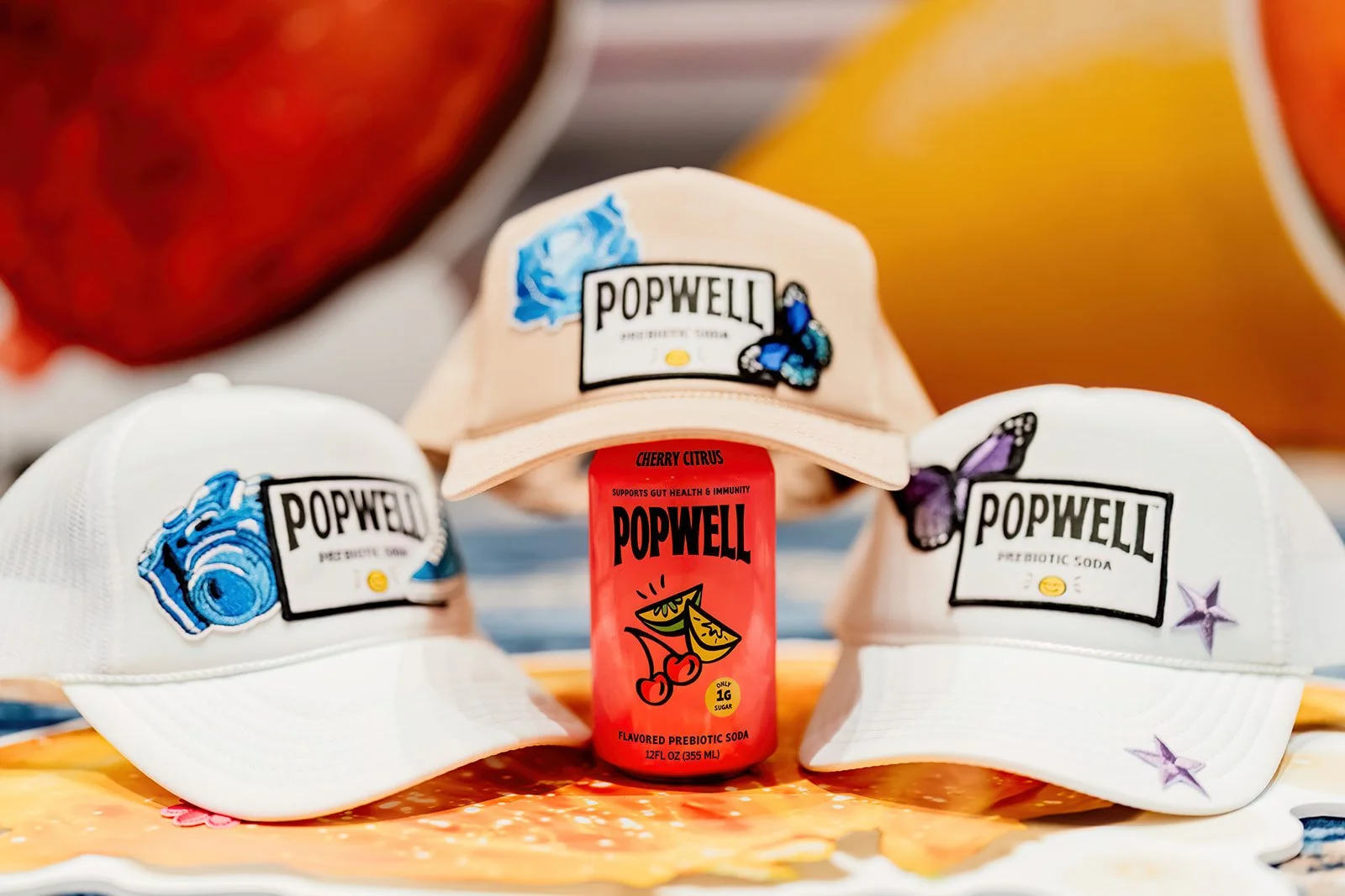 Three baseball caps with PopWELL branding and colorful patches like a blue wave, butterfly, and purple star. A small red bottle of PopWELL prebiotic soda is positioned in front, with a peach, orange, and a blurred red and yellow background.