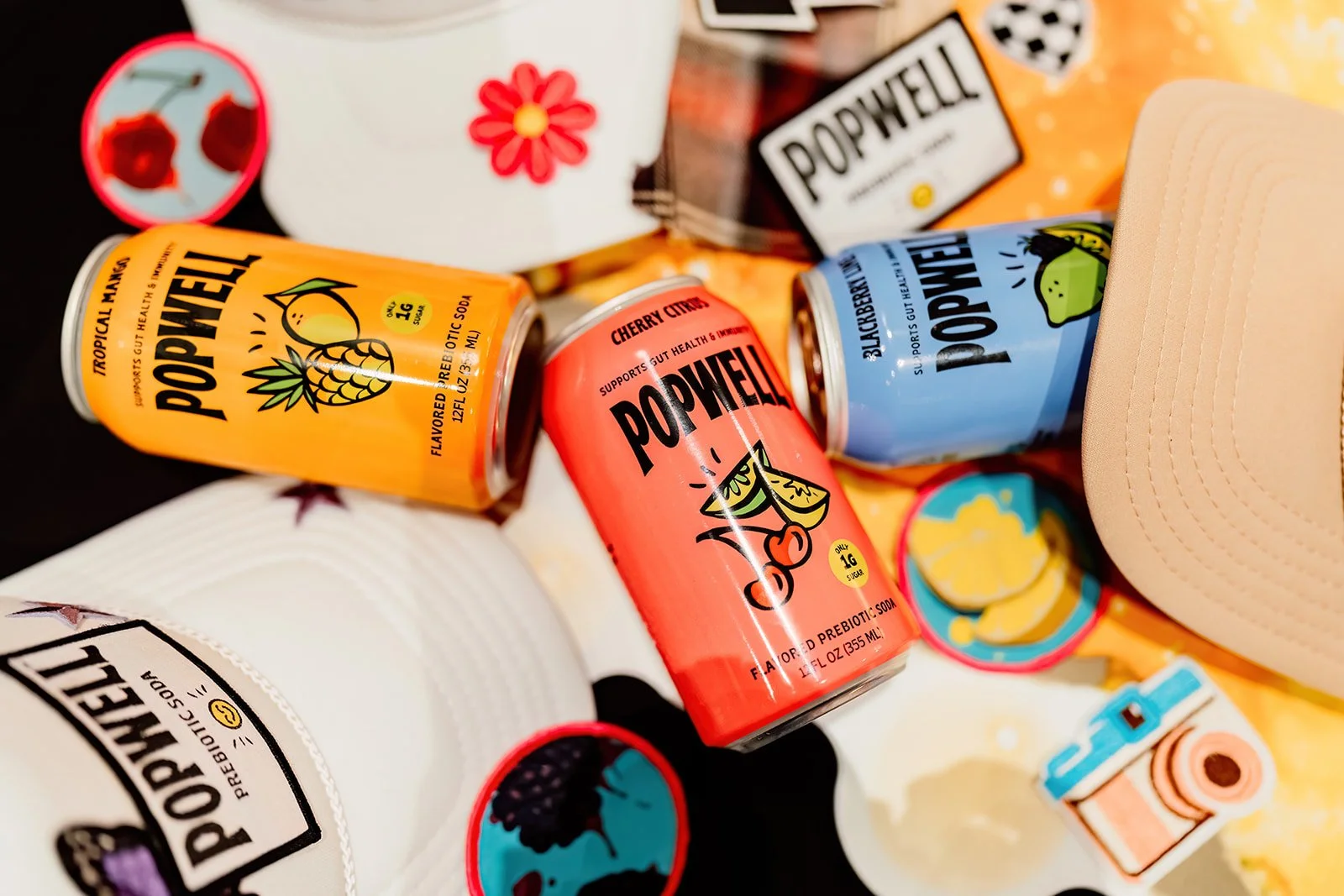 Colorful cans of Popwhel probiotic soda in various flavors, including pineapple and cherry citrus, surrounded by caps, a beige cap, and miscellaneous items on a table.