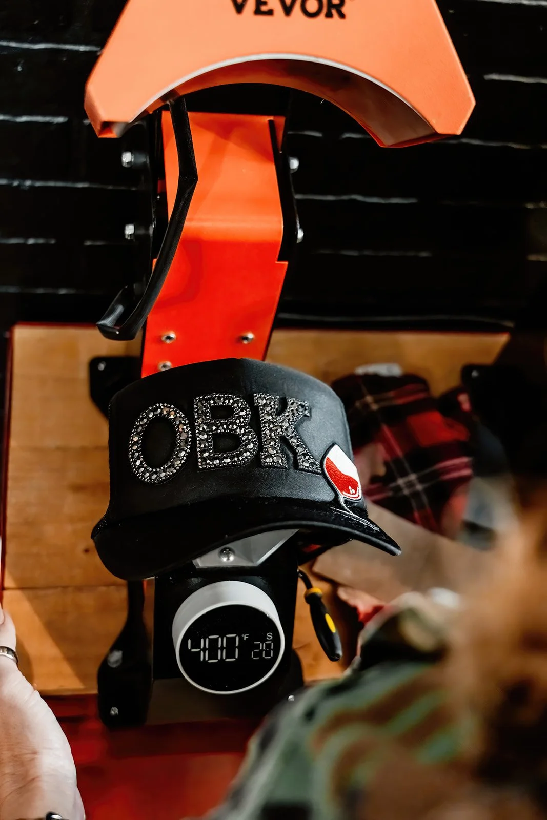 A black baseball cap with the word 'OBR' in rhinestones and a red, white, and black logo, placed on a digital temperature display showing 400°F. The display is on a wooden surface with a person wearing glasses and a plaid hat in the background.