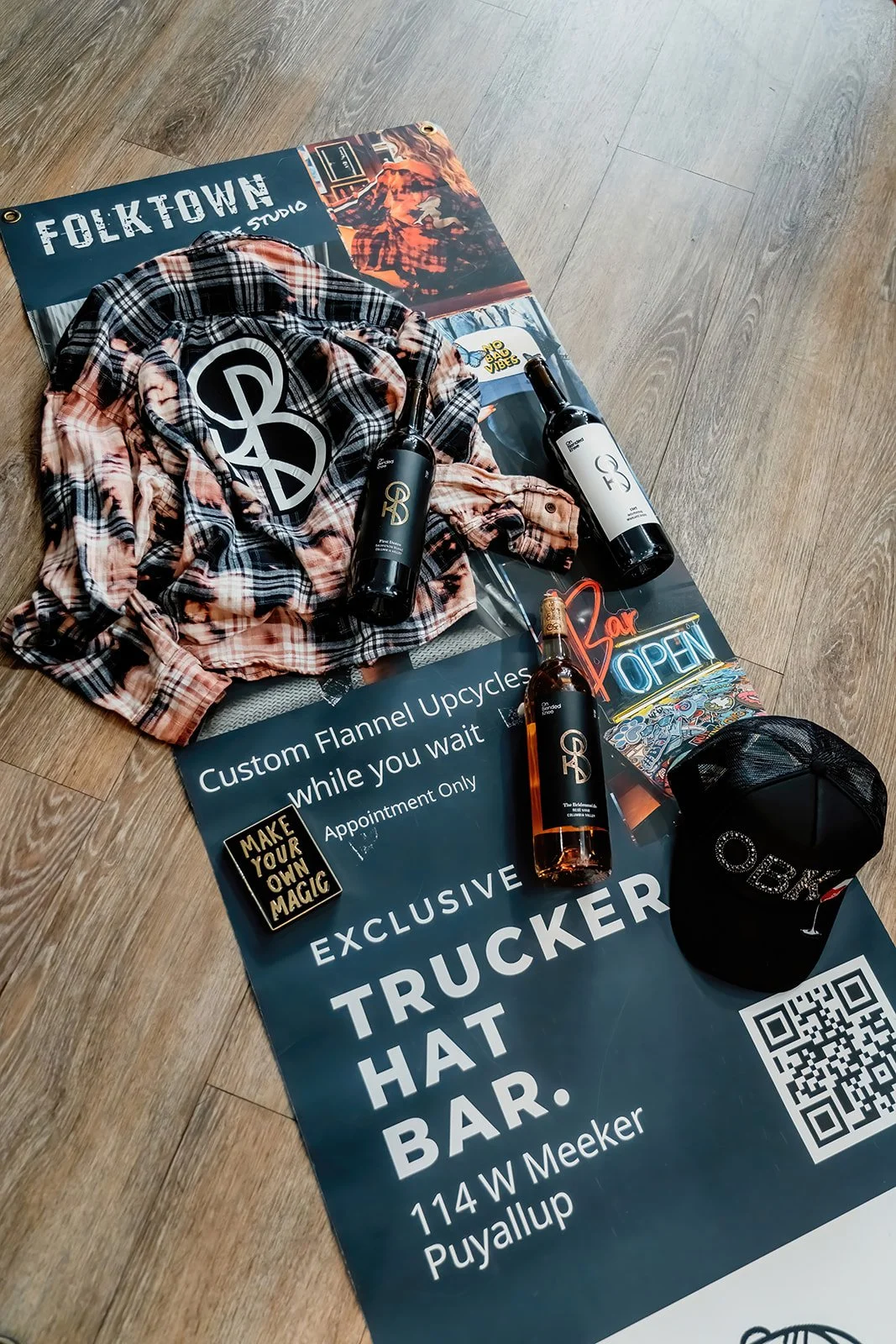 A promotional display for a trucker hat bar with bottles of wine, a cap, a plaid shirt with a peace sign patch, and various signs and stickers on a wood floor.
