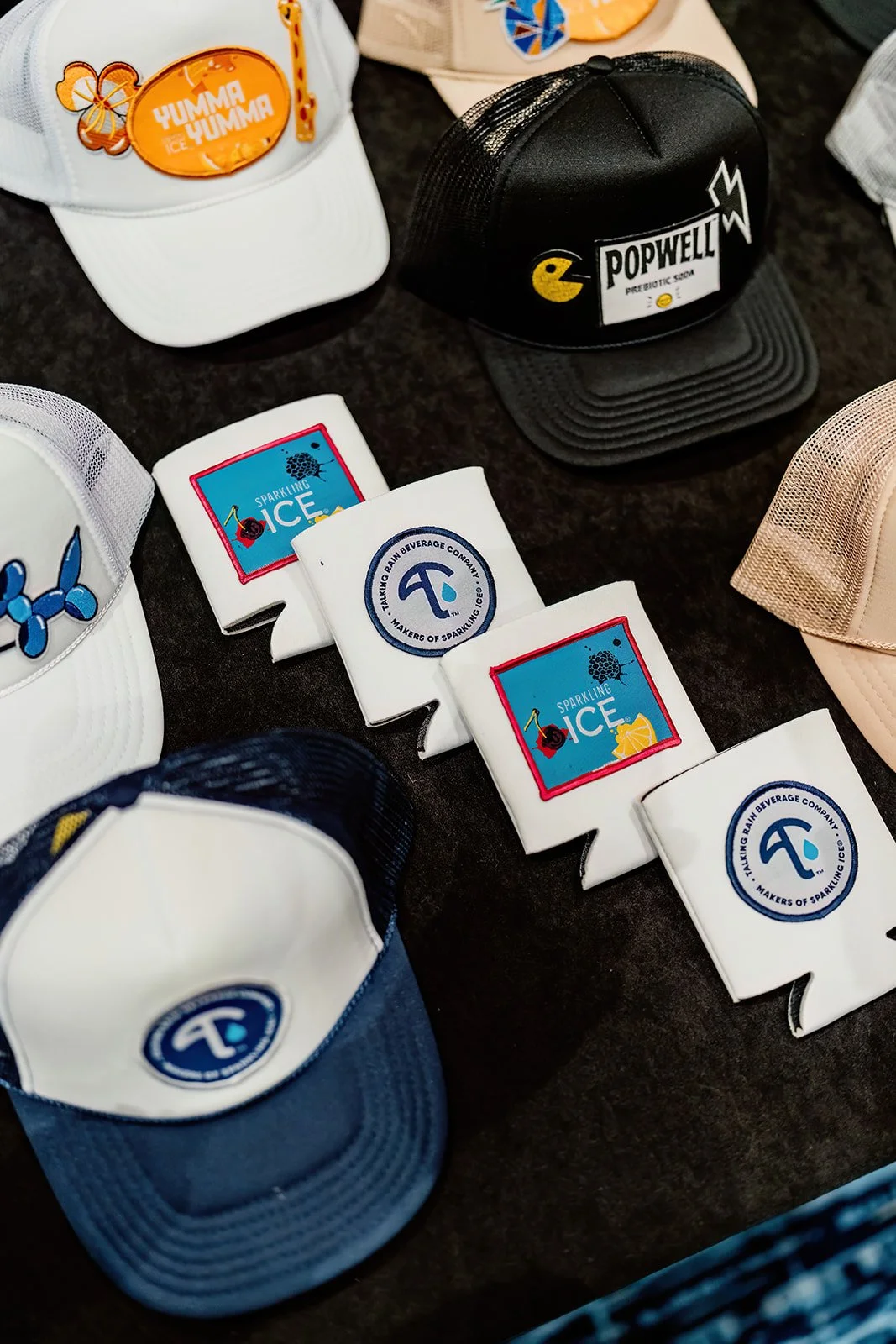 Various baseball caps and drink koozies with drink logos and designs displayed on a black surface.