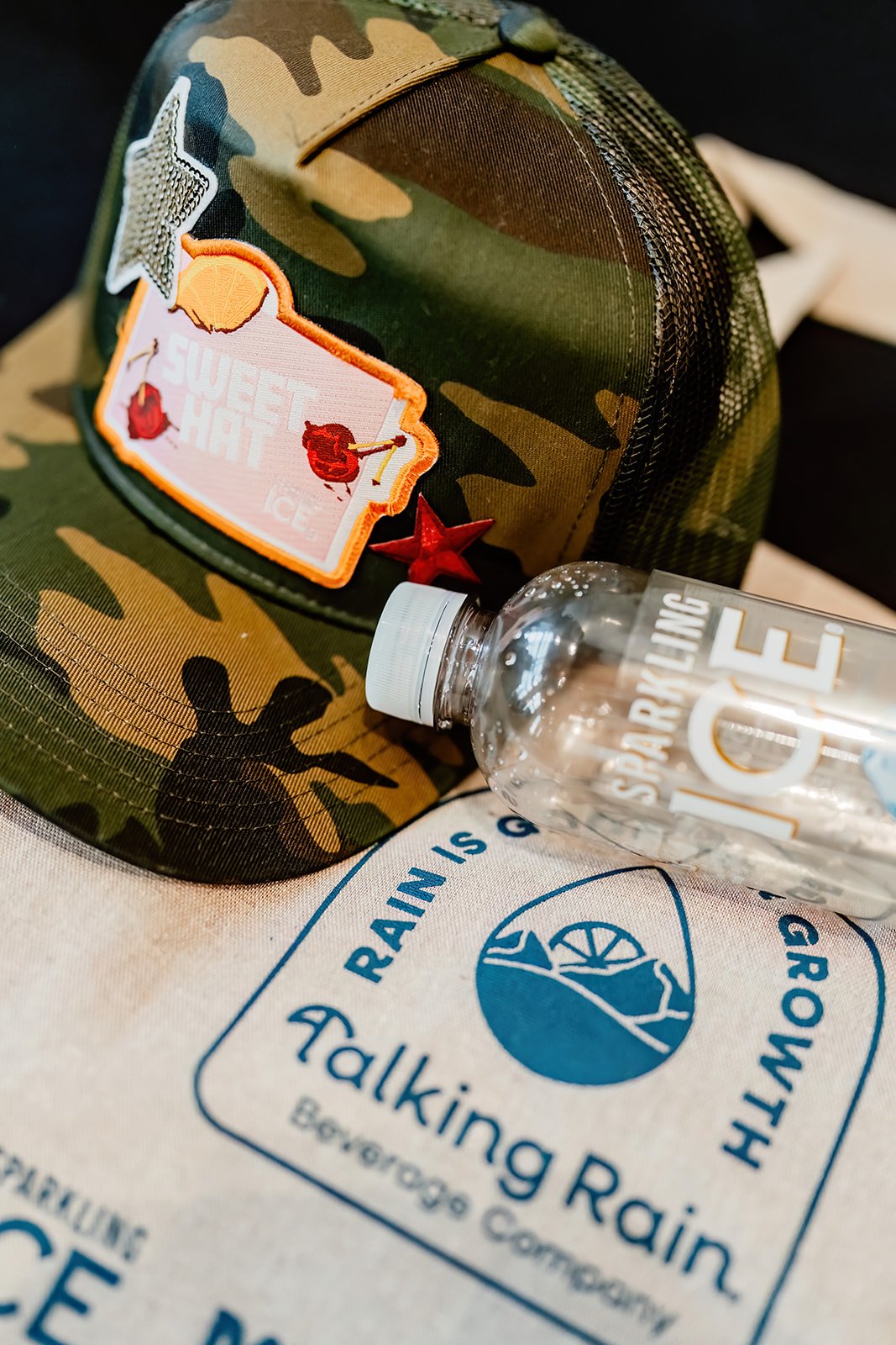 A camouflage baseball cap with patches, a clear water bottle, and a fabric bag with blue text and logo, placed on a black background.