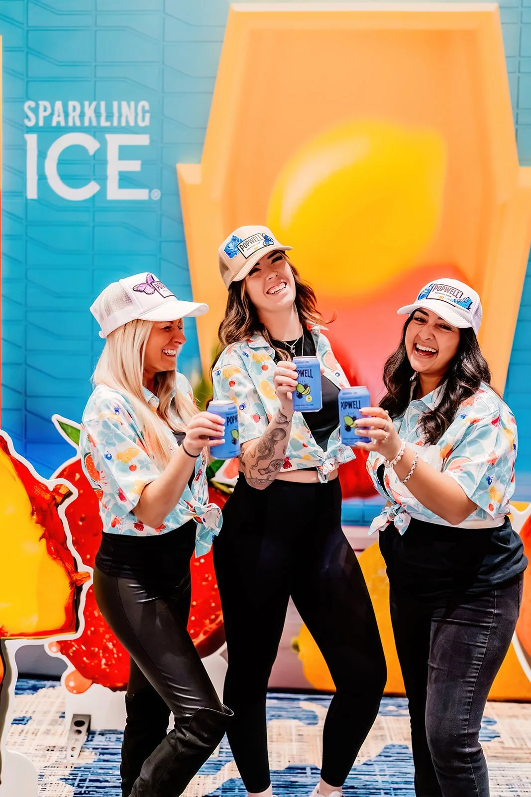 Three women wearing matching Hawaiian shirts and white caps, holding blue cans, standing in front of a colorful backdrop with the words 'Sparkling Ice' and images of fruit, smiling and posing for the photo.