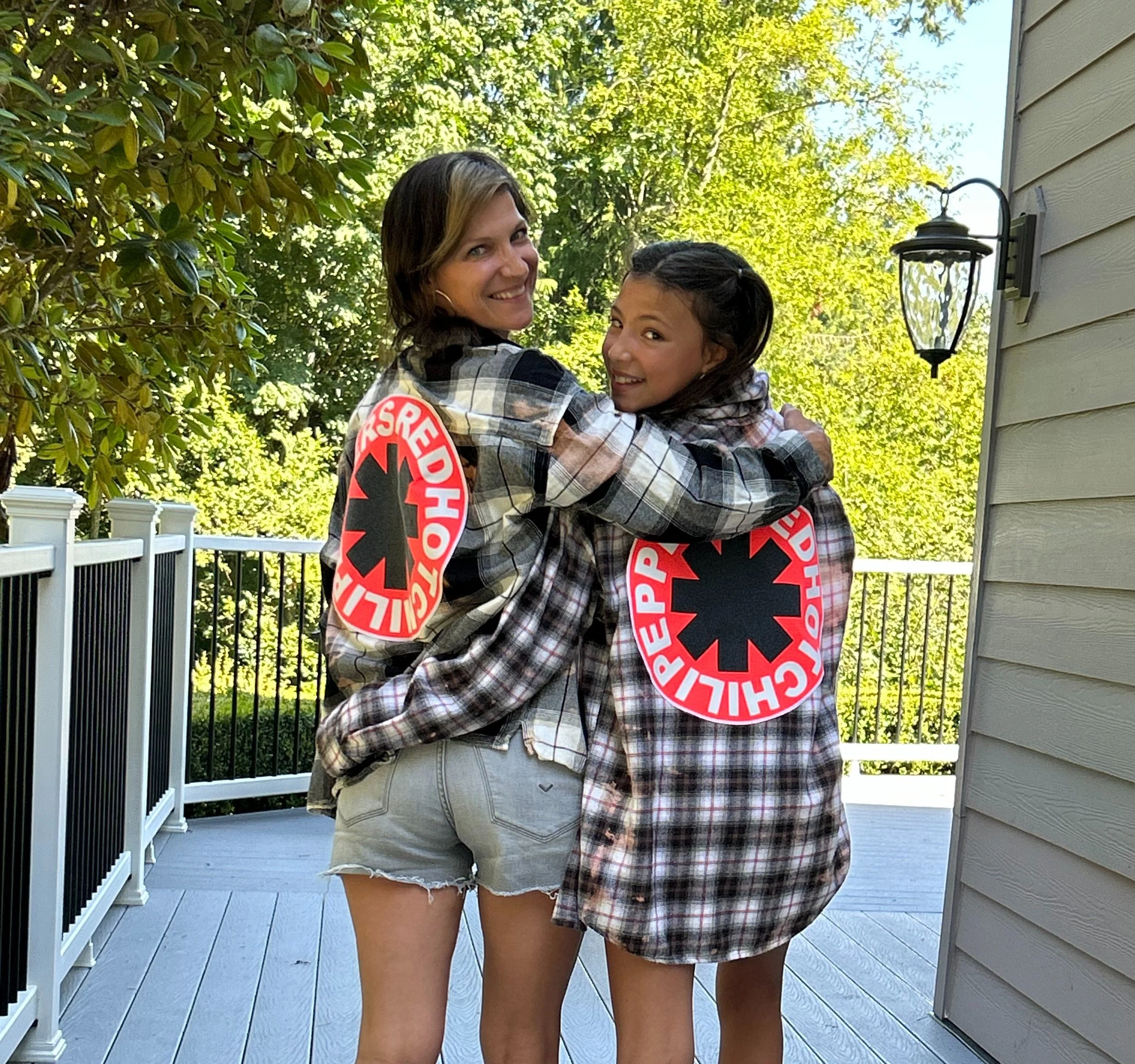 Two young women wearing plaid flannel shirts with Red Cross logos hugging and smiling on a porch with trees and a fence in the background.