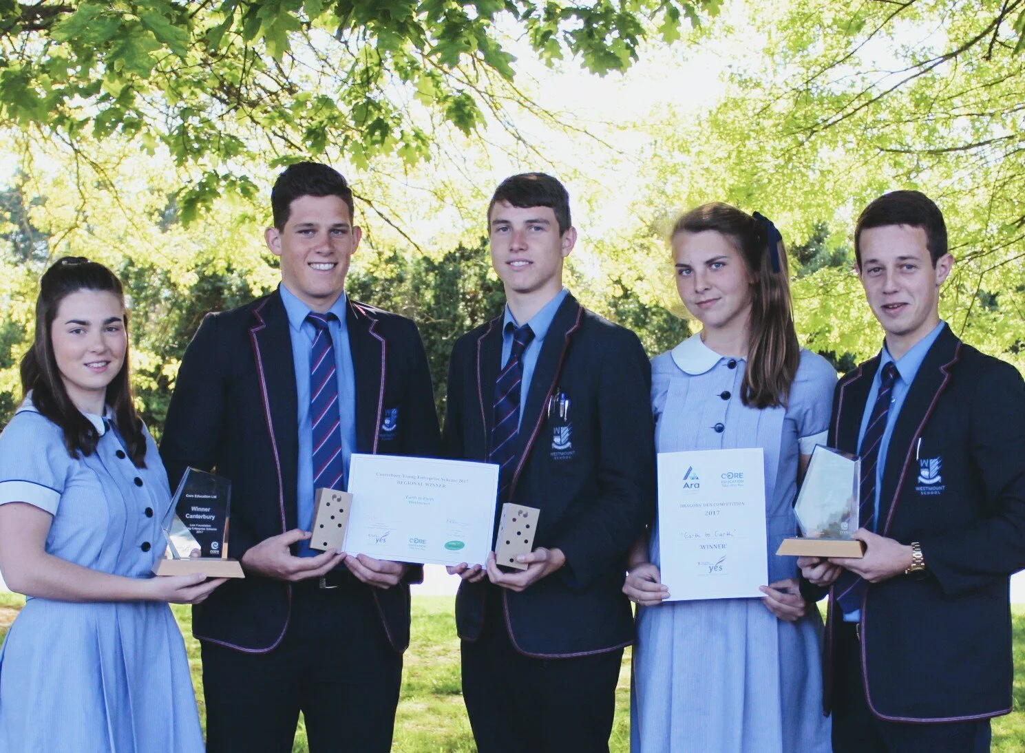 Five students dressed in school uniforms standing outdoors, holding awards and certificates, with lush green trees in the background.
