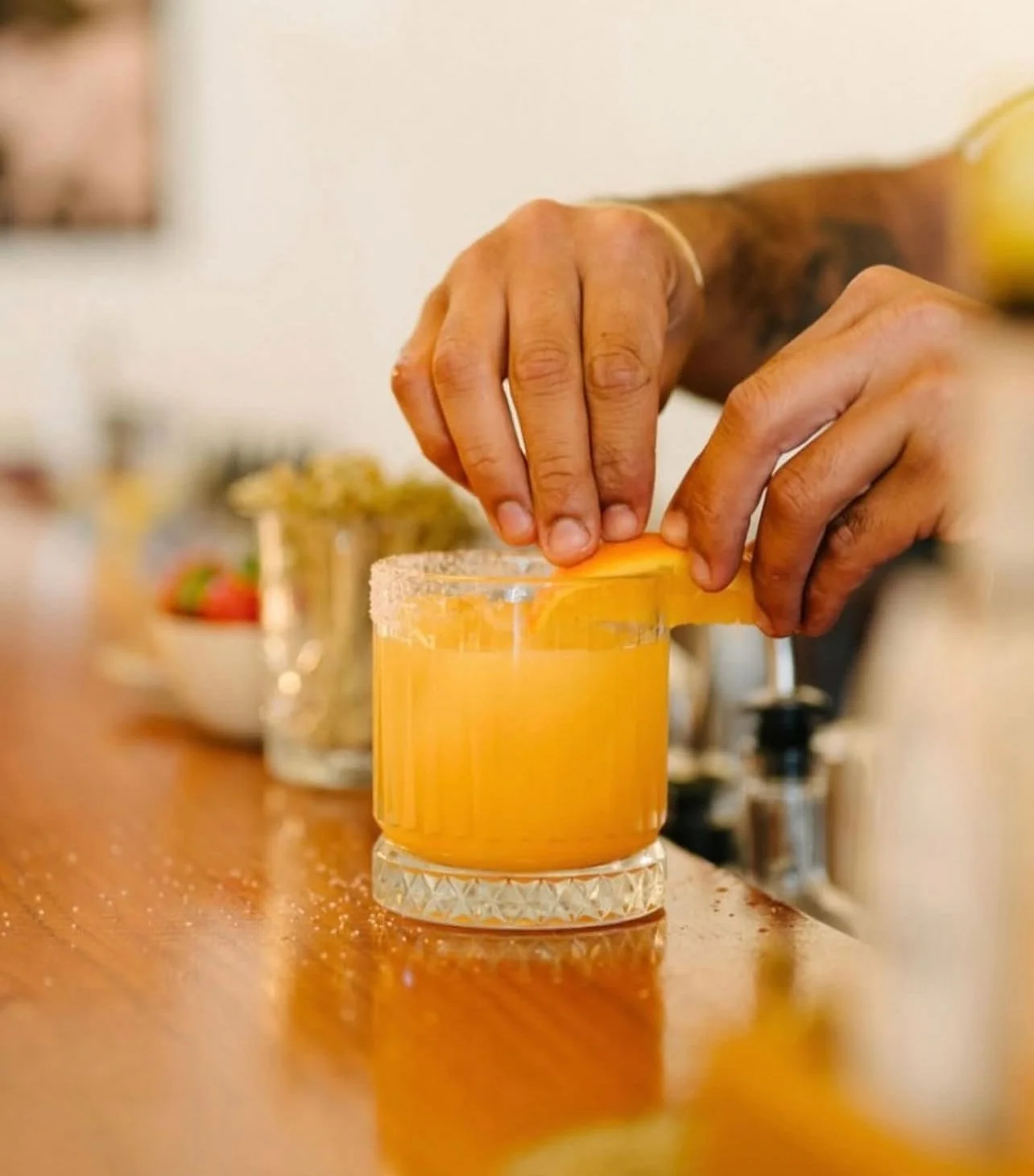 Person overlapping a glass of orange juice with a lemon wedge, salt rim, on a wooden bar counter.