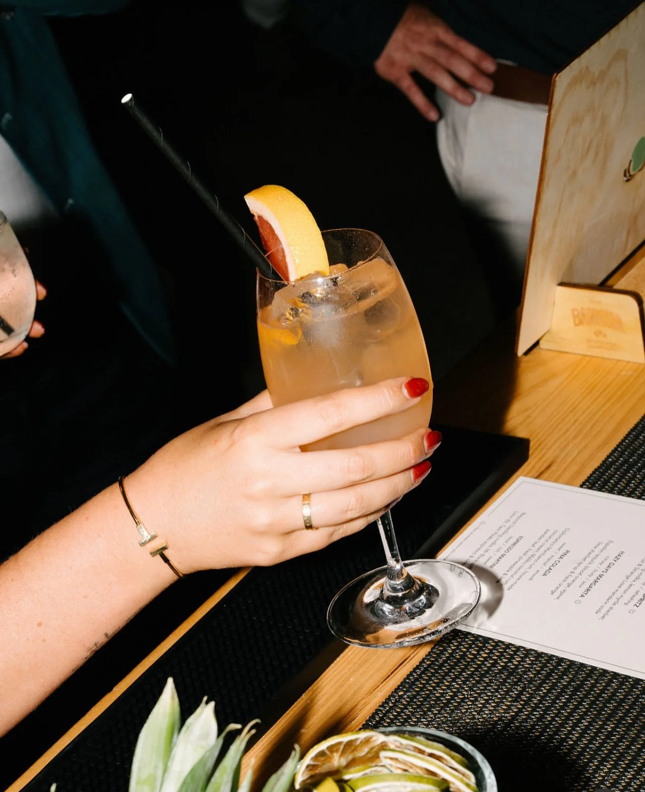 A woman's hand with red nail polish, wearing a ring and bracelet, holding a glass of cocktail garnished with an orange slice and a black straw, on a restaurant table.