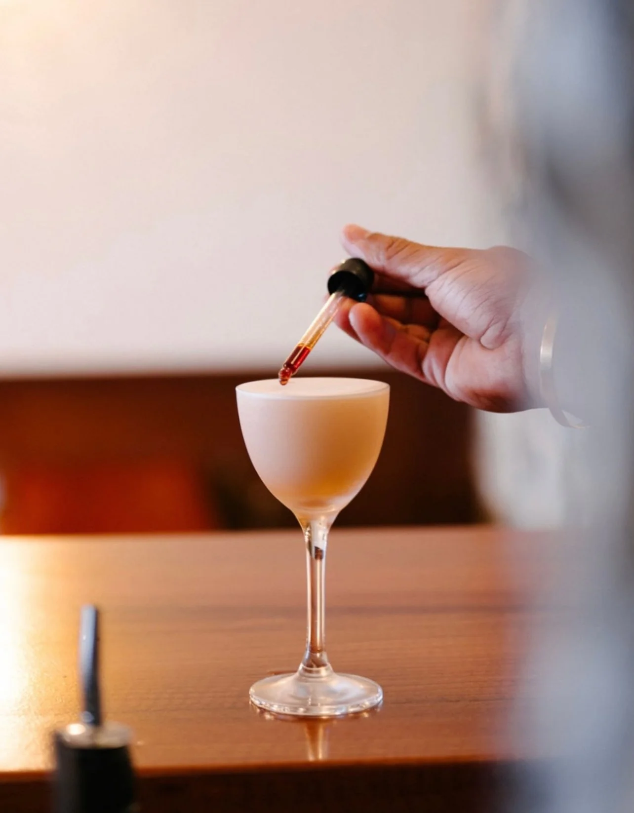 A person's hand holding a dropper filled with red liquid, adding it to a frosted cocktail glass on a wooden table.