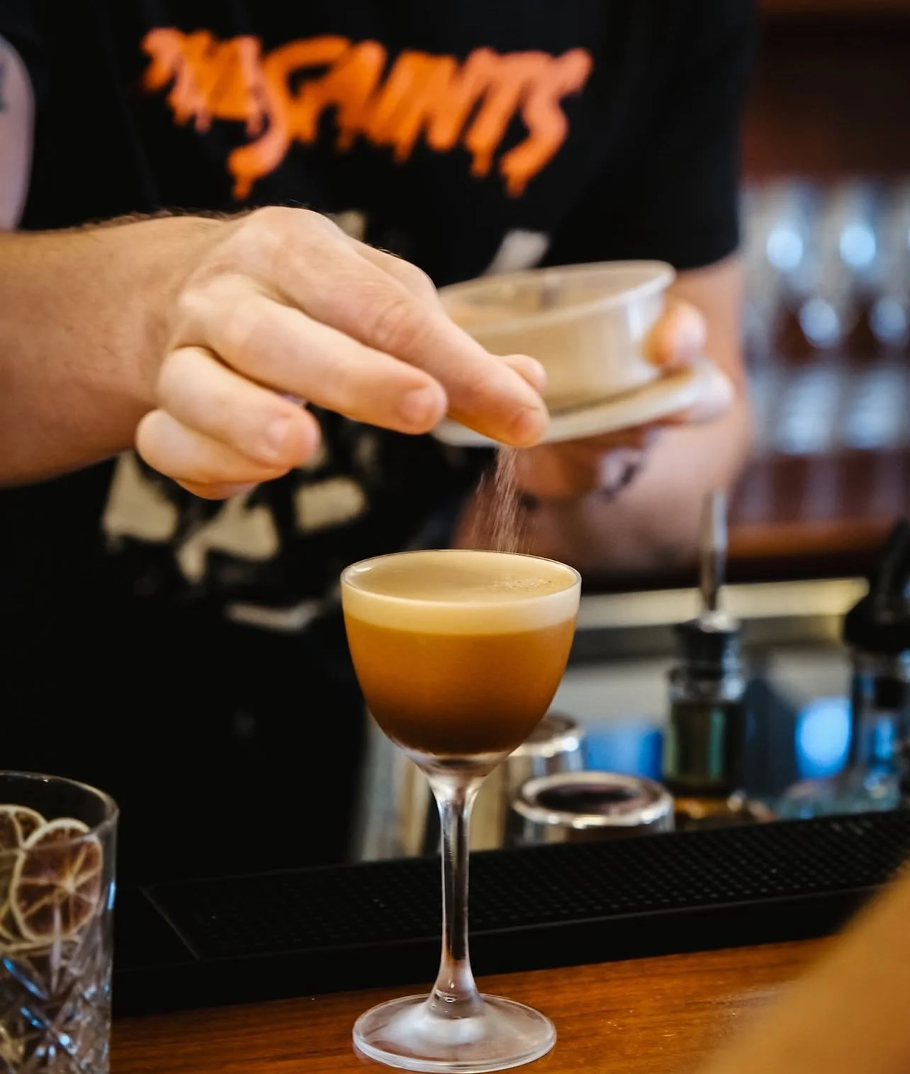A bartender pouring a powdered ingredient into a layered cocktail in a martini glass at a bar