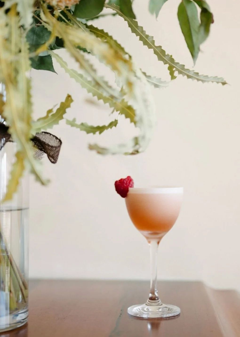 A cocktail in a martini glass garnished with a raspberry, placed on a wooden surface with a leafy plant in the background.